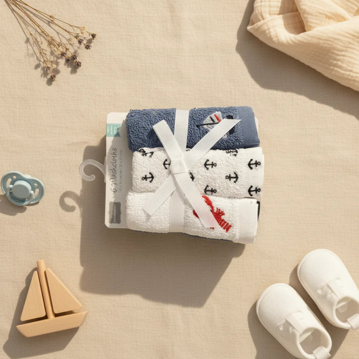 Nautical baby washcloths 6 pack flat lay from above on linen background showing navy blue anchor print and red lobster embroidery washcloths bundled with white ribbon bow with teal pacifier wooden sailboat toy white baby shoes and dried flowers