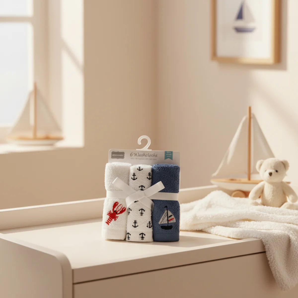 Nautical baby washcloths 6 pack in retail hanger packaging displayed on white dresser in warm cream nautical nursery room with wooden sailboat toy white teddy bear and framed sailboat wall art in background