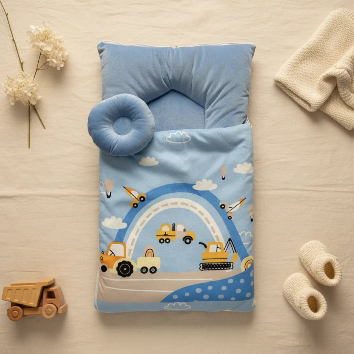 Construction vehicles baby sleeping bag flat lay from above on cream linen background showing open sky blue quilted sleeping bag with yellow dump trucks excavator cranes aeroplanes clouds and blue white rainbow arc print white polka dot lower border sky blue rectangular back pillow and round donut head pillow beside wooden dump truck toy white knit booties cream throw and dried white flowers