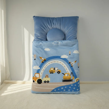 Construction vehicles baby sleeping bag studio scene propped upright against pale grey wall in natural light showing sky blue quilted sleeping bag with yellow dump trucks crawler excavator cranes aeroplanes hot air balloons and blue white rainbow arc on blue polka dot border with sky blue rectangular back pillow and round donut head pillow at top
