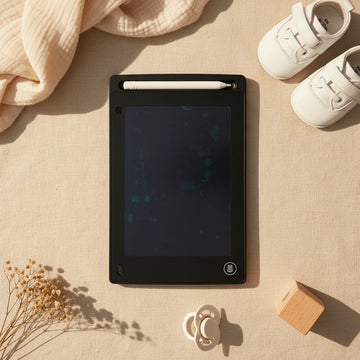 8.5 inch black LCD Writing Tablet flat lay showing sleek black housing with white stylus pen in top slot and single erase button on linen background with white baby shoes pacifier and wooden block