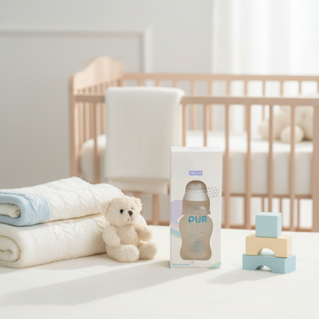 PUR Anti-Colic Baby Feeding Bottle BPA Free - cozy nursery scene in retail box on white table with teddy bear wooden blocks and natural wood crib in background