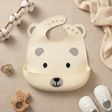 Cream bear face silicone baby bib with food catch pocket flat lay showing shaped bear design with grey ear accents cartoon eyes smiling bear face and snap button neck closure on linen background with wooden bear teether pacifier and white sneakers