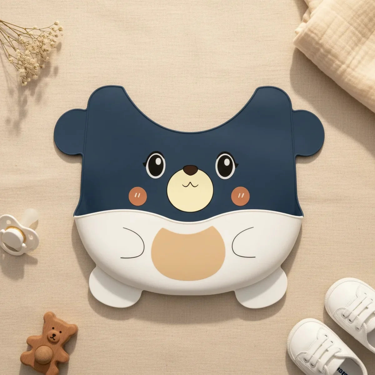 Dark blue bear face silicone baby bib flat lay showing full navy blue bear shaped bib with cream and tan bear face rosy cheeks big expressive eyes and white food catch pocket forming bear tummy on wood surface with baby bear teether white pacifier white sneakers and white gypsophila flowers