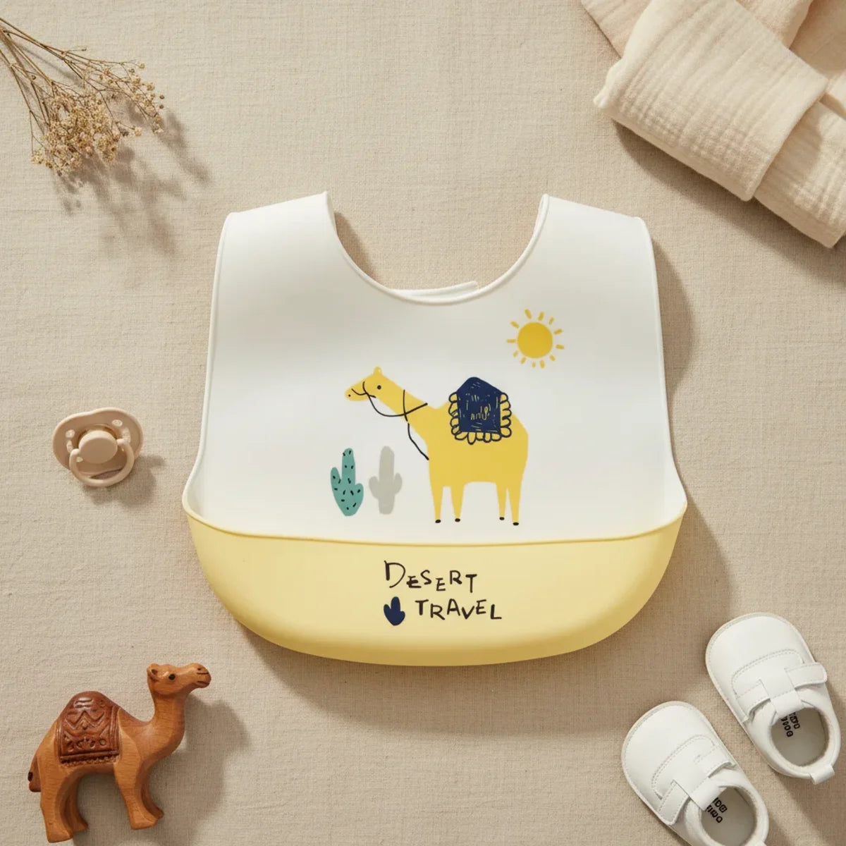 Desert Travel Camel silicone baby bib flat lay showing white bib with illustrated yellow camel navy blue saddle teal cactus and yellow sun scene with yellow food catch pocket stamped Desert Travel on linen background with ceramic camel figurine pacifier and white velcro sneakers