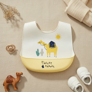Desert Travel Camel silicone baby bib flat lay showing white bib with illustrated yellow camel navy blue saddle teal cactus and yellow sun scene with yellow food catch pocket stamped Desert Travel on linen background with ceramic camel figurine pacifier and white velcro sneakers