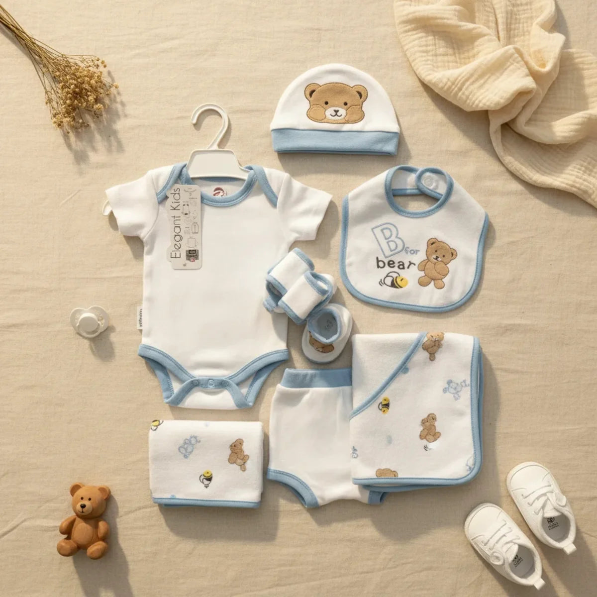 Elegant Kids B for Bear newborn gift set blue 7-piece flat lay showing white and blue trim bodysuit on hanger shorts bear hat B for bear bib booties swaddle blanket and muslin cloth on linen background with teddy bear and white sneakers