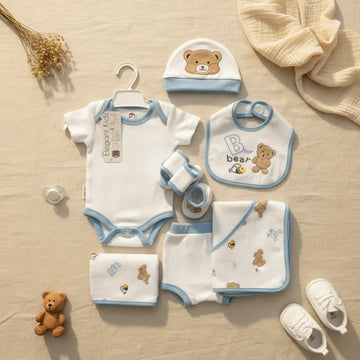 Elegant Kids B for Bear newborn gift set blue 7-piece flat lay showing white and blue trim bodysuit on hanger shorts bear hat B for bear bib booties swaddle blanket and muslin cloth on linen background with teddy bear and white sneakers