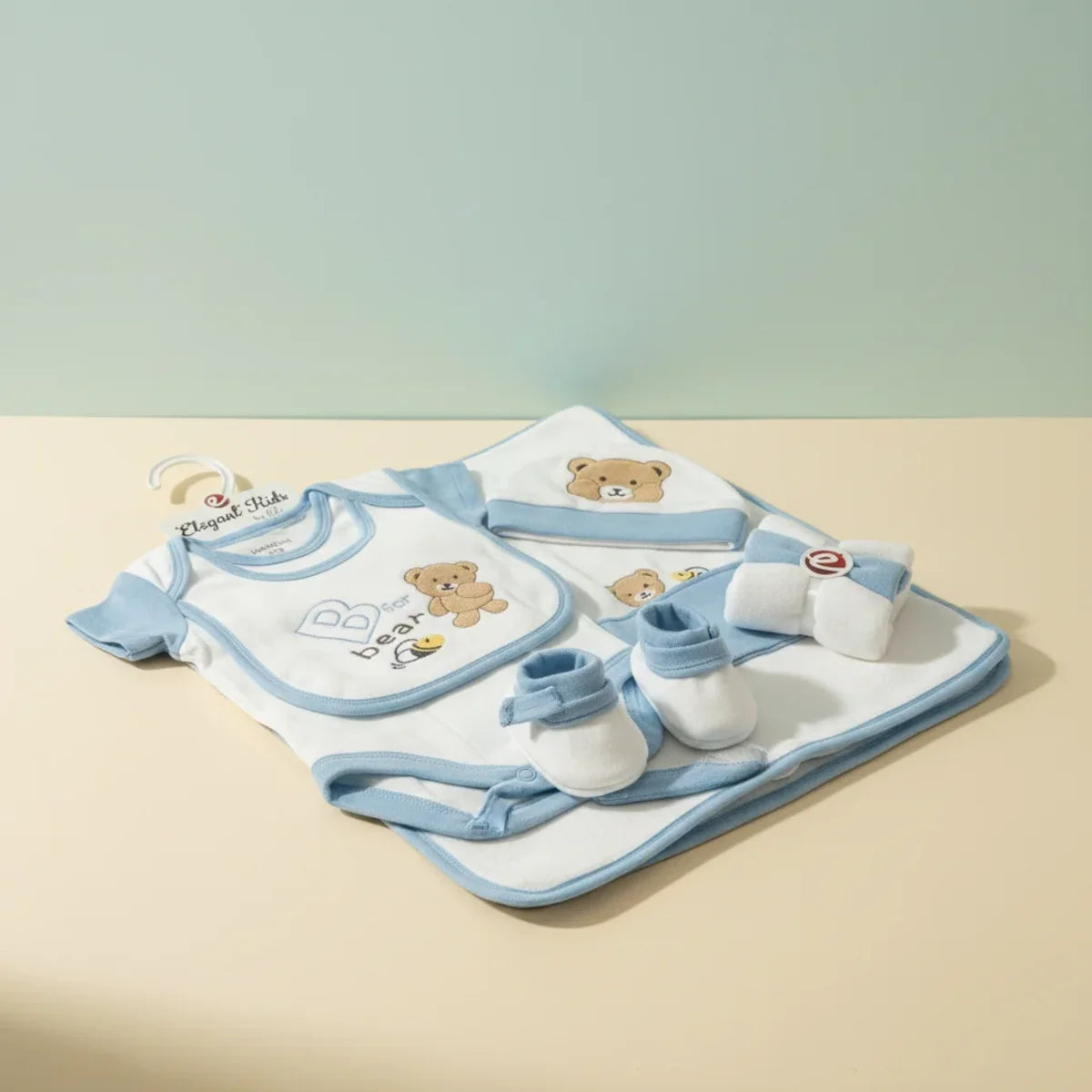 Elegant Kids B for Bear newborn gift set blue studio scene showing bodysuit bib hat booties and blanket laid on sage green and cream studio background with Elegant Kids branded hanger tag