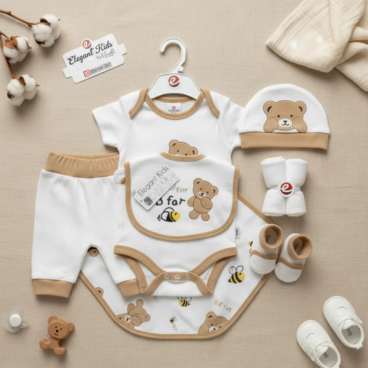 Elegant Kids B for Bear newborn gift set brown camel 10-piece starter set flat lay showing white and camel trim bodysuit on hanger trousers bear hat bib booties and muslin cloth on linen background with cotton stems and white sneakers