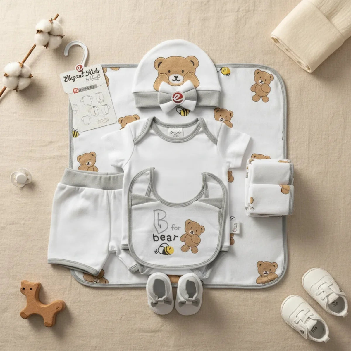 Elegant Kids B for Bear newborn gift set grey 10-piece starter set flat lay showing all-over bear bee print blanket bodysuit shorts bear print hat bib bow headband grey booties and muslin cloths on linen background with wooden horse and white sneakers