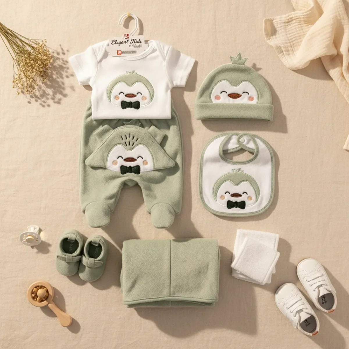 Elegant Kids Penguin Newborn Gift Set sage green 7-piece flat lay showing bodysuit footed trousers penguin hat bib booties blanket and muslin cloth on linen background with wooden rattle pacifier and white sneakers