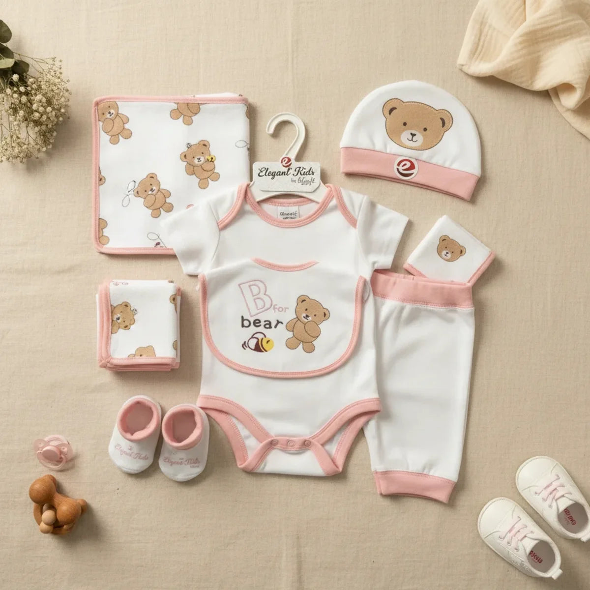Elegant Kids B for Bear newborn gift set pink 7-piece flat lay showing white and blush pink trim bodysuit leggings bear hat bib booties all-over bear print blanket and muslin cloths on linen background with wooden teether and white sneakers
