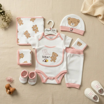 Elegant Kids B for Bear newborn gift set pink 7-piece flat lay showing white and blush pink trim bodysuit leggings bear hat bib booties all-over bear print blanket and muslin cloths on linen background with wooden teether and white sneakers