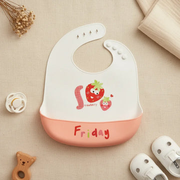 Friday Strawberry silicone baby bib flat lay showing white bib with illustrated smiling strawberry characters and Strawberry text with soft peach food catch pocket stamped Friday in red on linen background with wooden bear teether white pacifier and white sandals