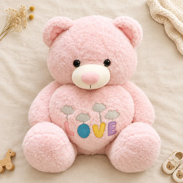 Giant pink LOVE teddy bear plush toy flat lay from above on cream linen background showing full front view of oversized curly boucle pink teddy bear with cream white muzzle dome shiny pink nose glossy black bead eyes soft pink inner ears and tummy with four colourful felt LOVE letter appliques in pink teal yellow and lavender each suspended on embroidered thread from grey embroidered cloud beside dried yellow billy balls flowers cream knit blanket and white baby shoes