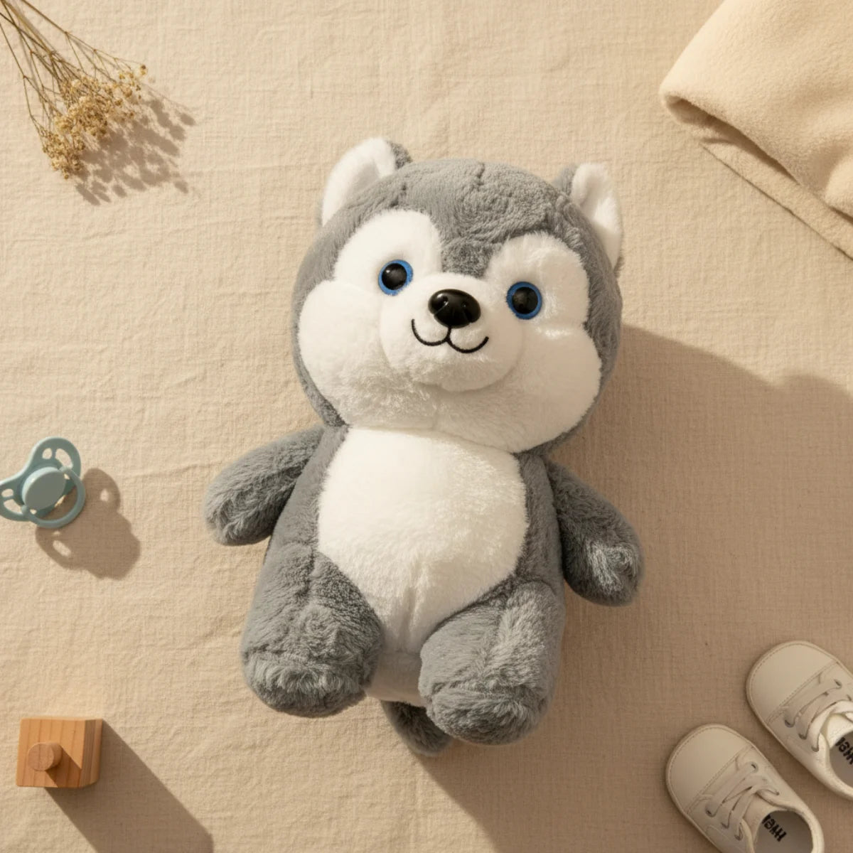 Baby husky plush toy flat lay from above on linen background showing full front face of grey white chubby husky puppy plush with sparkling blue eyes black nose and embroidered smile beside teal blue pacifier wooden block toy white baby sneakers and dried grass stems with cream knit throw corner