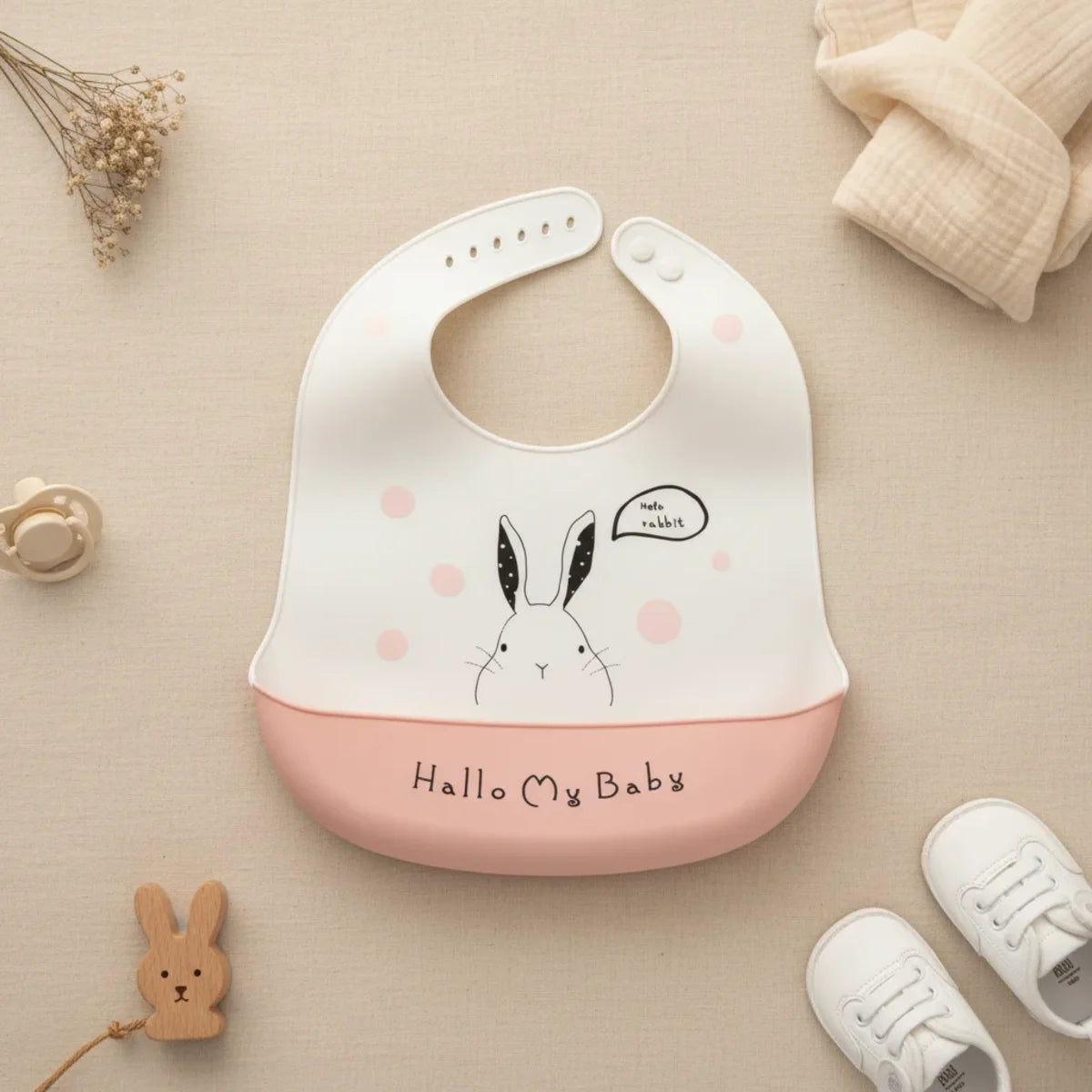 Hallo My Baby pink rabbit silicone baby bib flat lay showing white bib with minimalist line-drawn rabbit illustration Hello Rabbit speech bubble blush pink polka dots and pink food catch pocket stamped Hallo My Baby on linen background with wooden rabbit teether white pacifier and white lace-up sneakers