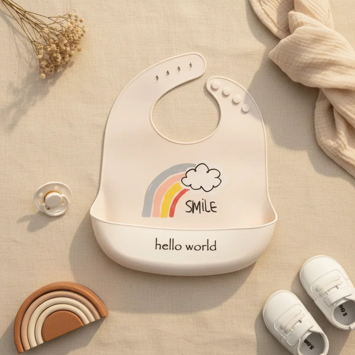 Hello World rainbow smile silicone baby bib flat lay showing cream bib with pastel boho rainbow cloud and SMILE illustration and cream food catch pocket stamped hello world on linen background with wooden rainbow stacker toy white pacifier and white velcro sneakers