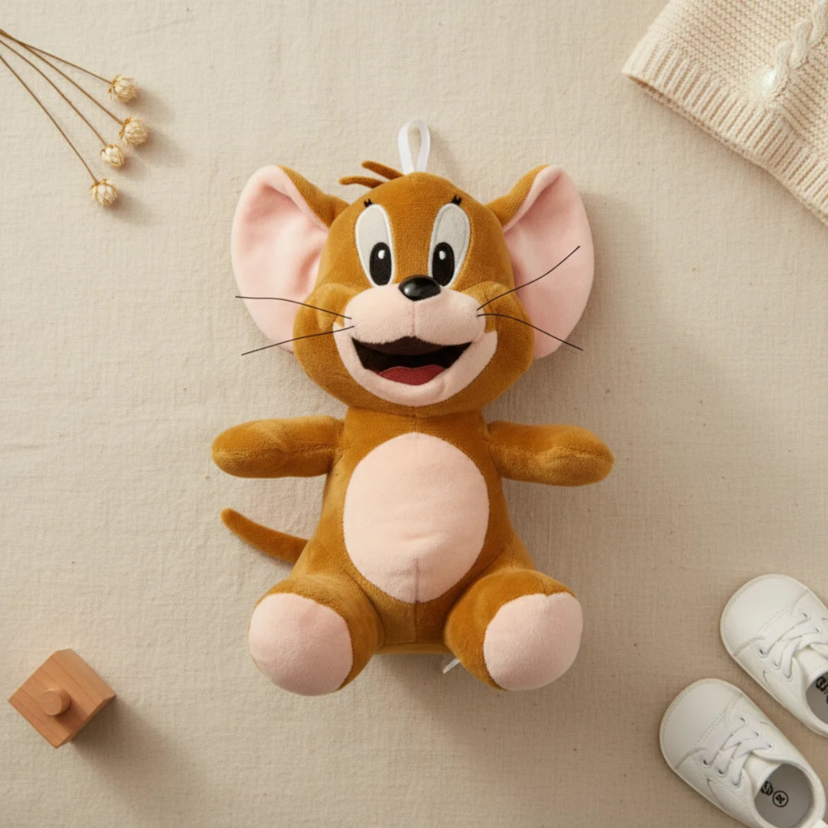 Jerry Mouse plush toy flat lay from above on cream linen background showing full golden brown Tom and Jerry cartoon mouse plush with enormous pink inner ears wide open-mouthed smile silver thread whiskers cream belly pink paw pads and long thin tail in classic arms-out sitting pose beside white mini sneakers dried cotton stems and cream knit throw