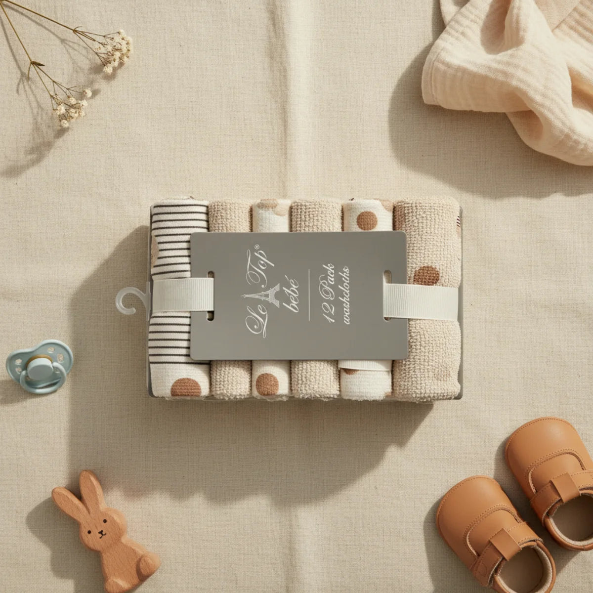 Le Top Bebe baby washcloths 12 pack flat lay from above on linen background showing grey Le Top bebe hanger card with 12 rolled terry washcloths in black pinstripe warm beige dot print and solid oat cream patterns with blue pacifier wooden bunny toy tan leather baby sandals and dried white flowers