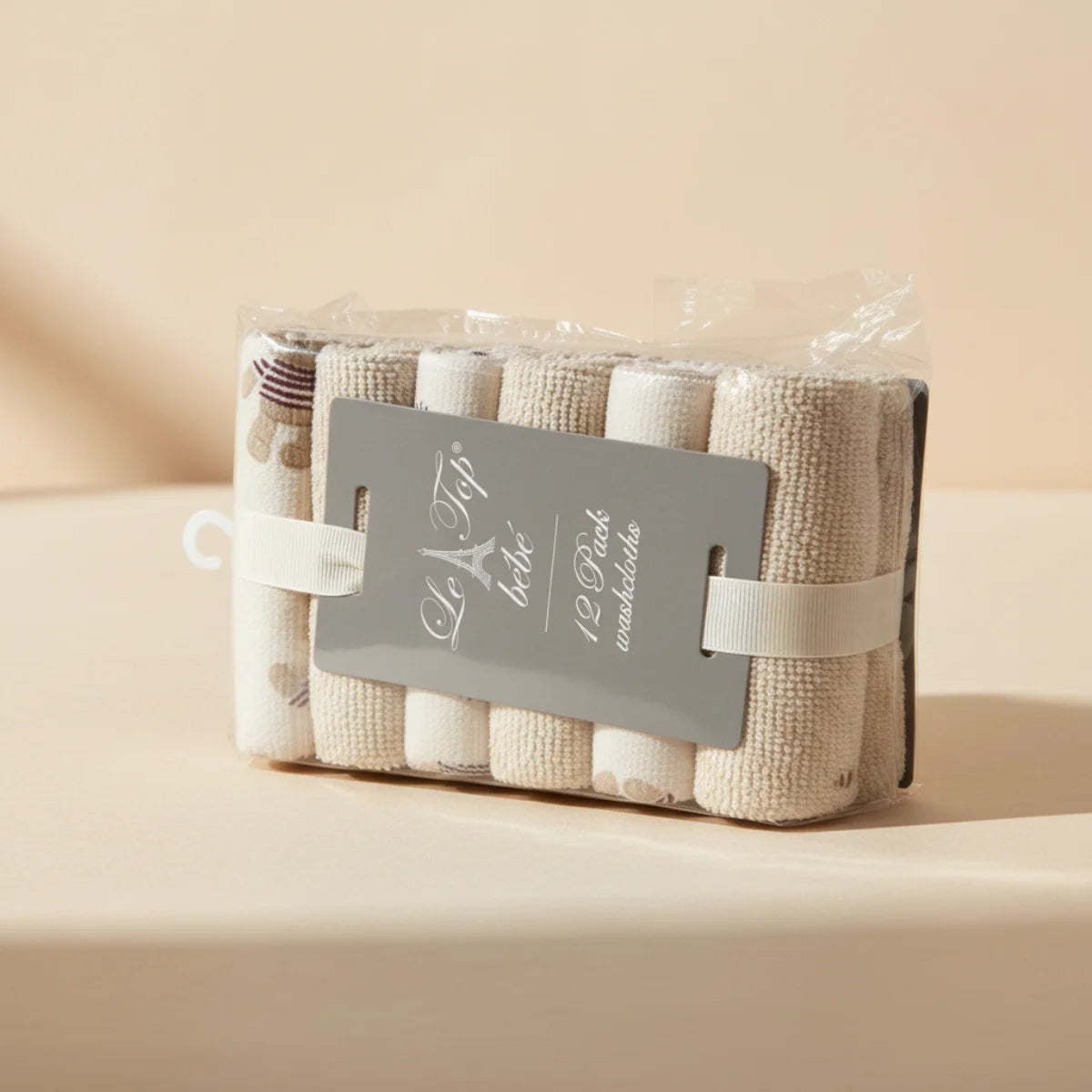 Le Top Bebe baby washcloths 12 pack in clear polybag with grey hanger card studio shot on warm cream beige background showing all 12 rolled terry washcloths in black stripe beige dot and solid oat cream patterns in transparent packaging