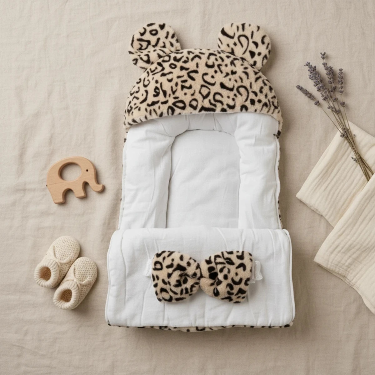 Leopard Bear-Ear Baby Nest flat lay open showing soft white quilted interior bear ears hood and matching leopard bow pillow with baby booties wooden elephant toy and dried lavender on linen background