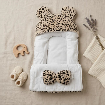 Leopard Bear-Ear Baby Nest flat lay open showing soft white quilted interior bear ears hood and matching leopard bow pillow with baby booties wooden elephant toy and dried lavender on linen background