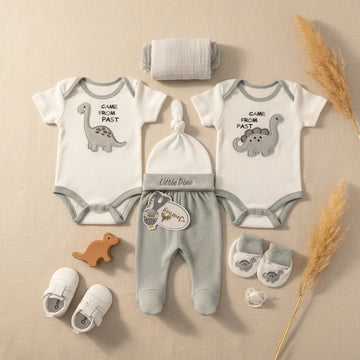 Little Dino Newborn Gift Set grey flat lay showing two Came From Past dinosaur bodysuits footed trousers knotted hat mittens booties and muslin on beige background with pampas grass and wooden dinosaur toy