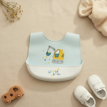 Little Star excavator silicone baby bib flat lay showing soft sky blue bib with illustrated yellow excavator grey arm teal bucket tracked wheels and star sparkle details with white food catch pocket stamped Little Star in blue and yellow on linen background with wooden car toy and white velcro sneakers