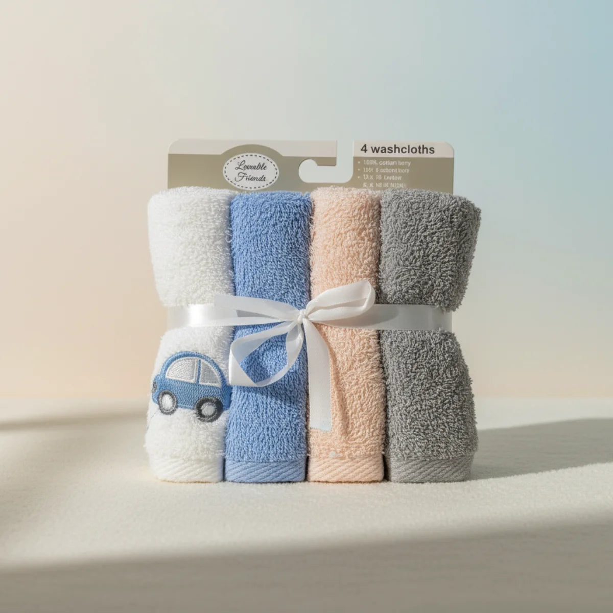 Lovable Friends baby washcloth set of 4 studio scene on soft warm cream gradient background showing four rolled cotton terry washcloths in white sky blue blush pink and grey tied with white satin ribbon bow with embroidered navy car applique in retail packaging with soft natural shadow