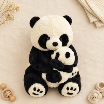 Mama and baby panda plush toy flat lay from above on cream linen background showing large mama giant panda in bold black white plush with deep jet-black eye patches glossy eyes shiny nose round black ears and embroidered toe paw pads cradling tiny miniature baby panda cub in same design with tiny glossy eyes and embroidered paw pads beside wooden teether ring cream knit blanket bear baby shoes and dried white flowers
