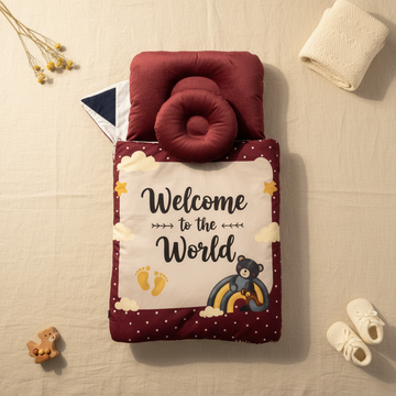 Welcome to the World baby sleeping bag flat lay open from above on cream background showing deep maroon donut head pillow rich burgundy quilted exterior white polka dot border navy white interior lining and illustrated white panel with navy bear on rainbow gold stars yellow footprints and fluffy clouds beside dried flowers and cream knit throw