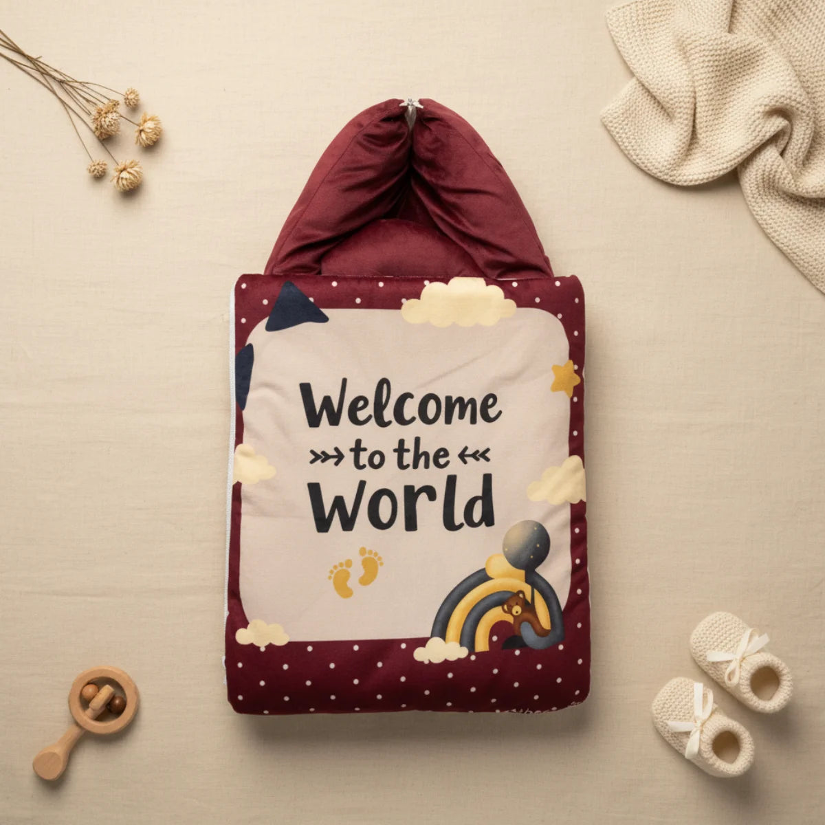Welcome to the World baby sleeping bag closed flat lay from above on cream linen background showing full maroon quilted sleeping bag with white polka dot border and illustrated central panel with Welcome to the World text navy bear rainbow gold stars yellow footprints and clouds with burgundy zip closure top beside dried flowers cream throw and white baby booties