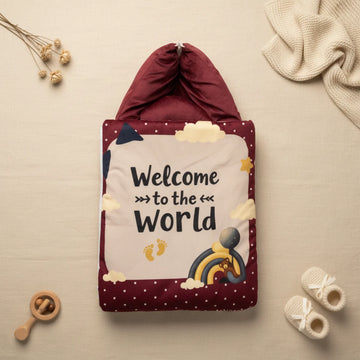 Welcome to the World baby sleeping bag closed flat lay from above on cream linen background showing full maroon quilted sleeping bag with white polka dot border and illustrated central panel with Welcome to the World text navy bear rainbow gold stars yellow footprints and clouds with burgundy zip closure top beside dried flowers cream throw and white baby booties