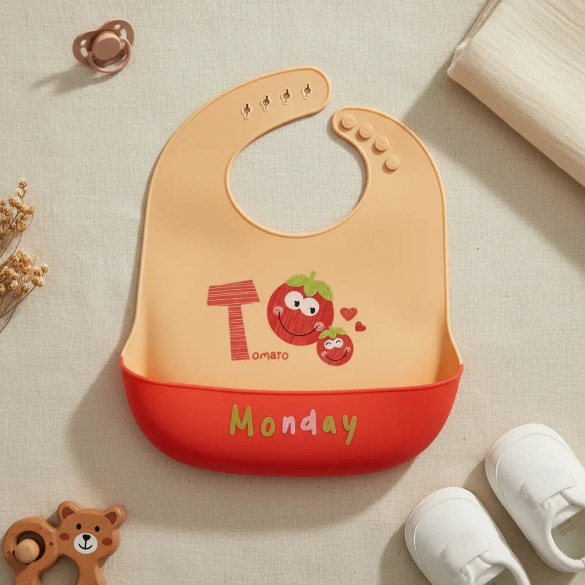 Monday Tomato silicone baby bib flat lay showing warm peach bib with illustrated smiling tomato characters bold T letter and Tomato text with deep red food catch pocket stamped Monday in green lettering on linen background with wooden bear ring teether brown pacifier and white sneakers