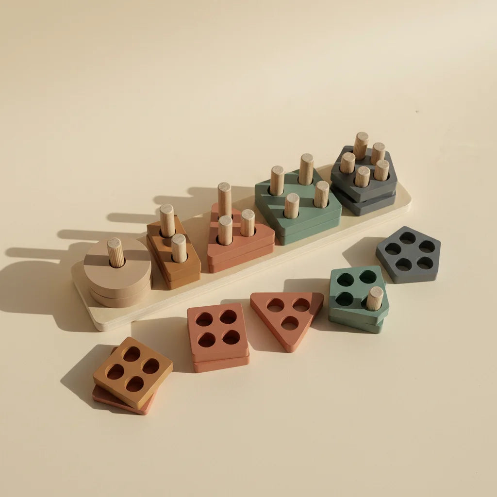 Montessori geometric shape sorter stacker studio shot showing 5 peg wooden board with sand cream terracotta rust sage green and slate grey geometric shape stacks with loose square triangle and pentagon pieces scattered on warm beige background