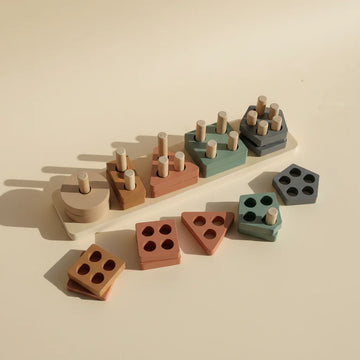 Montessori geometric shape sorter stacker studio shot showing 5 peg wooden board with sand cream terracotta rust sage green and slate grey geometric shape stacks with loose square triangle and pentagon pieces scattered on warm beige background