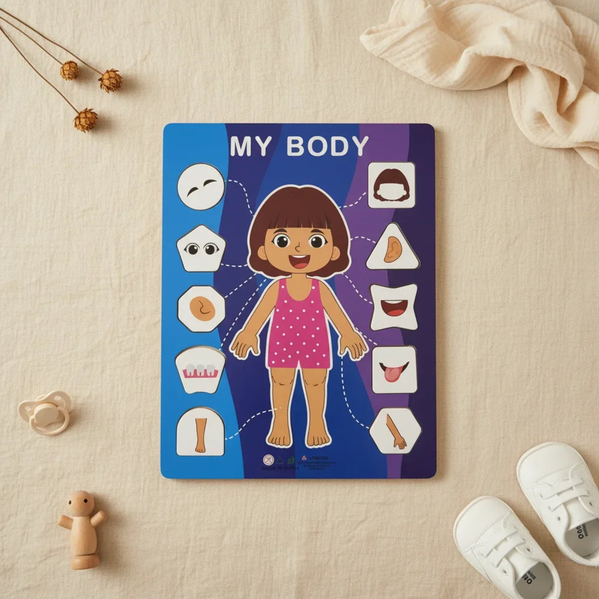 My Body wooden puzzle board girl version flat lay on linen background showing blue purple gradient board with illustrated smiling girl pink polka dot outfit and 10 shaped lift-out body part pieces with wooden figurine pacifier and white baby sneakers dried flowers