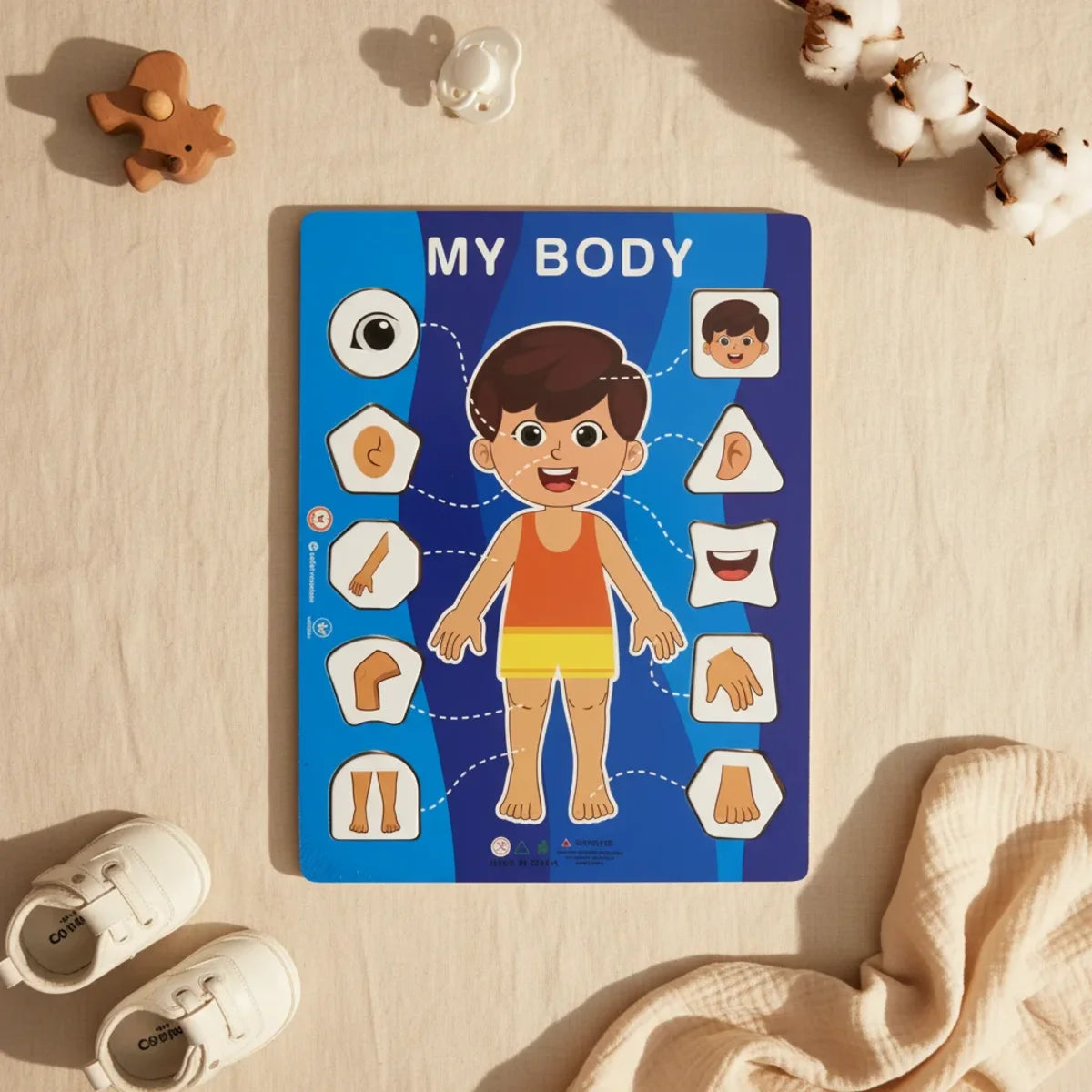 My Body wooden puzzle board boy version flat lay on linen background showing blue board with illustrated smiling boy orange yellow outfit and 10 shaped lift-out body part pieces eye ear nose mouth hand feet arm with wooden bear figurine pacifier and white baby sneakers