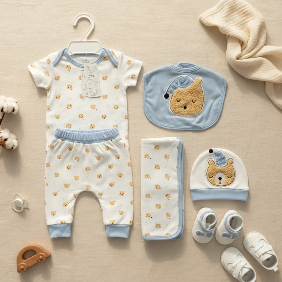 New Journey Sleeping Bear newborn gift set blue 5-piece flat lay showing white and sky blue trim all-over sleeping bear print bodysuit on hanger matching trousers 3D bear hat 3D bear bib bear print blanket on linen background with wooden car teether and white sneakers