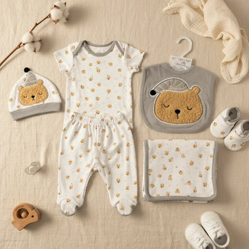 New Journey Sleeping Bear newborn gift set grey 5-piece flat lay showing white and soft grey trim all-over sleeping bear print bodysuit footed leggings 3D bear hat grey 3D bear bib and all-over bear print blanket on linen background with wooden elephant teether and white sneakers