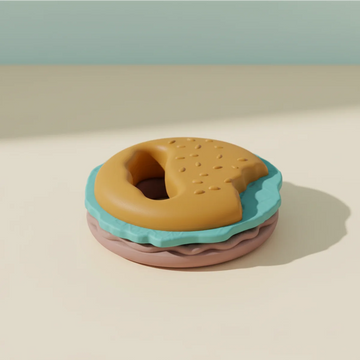 Burger silicone baby teether studio scene showing golden sesame bun top with textured dots and ergonomic ring handle teal wavy lettuce layer and mauve pink bun base stacked on warm cream background with soft teal shadow