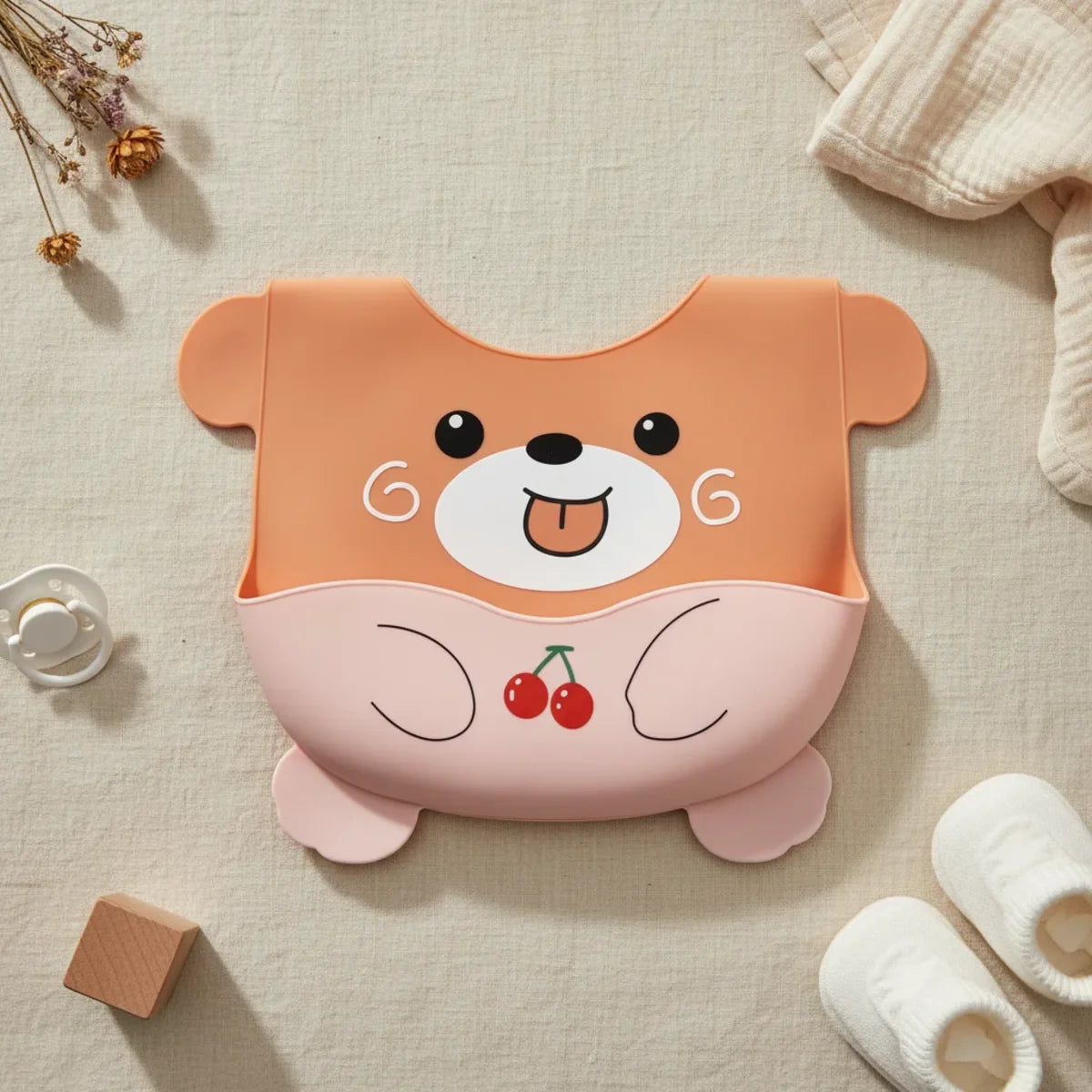 Orange bear cherry silicone baby bib flat lay showing full burnt orange bear shaped bib with round ears chubby arms bear feet expressive face tongue out and blush pink tummy pocket with red cherry illustration on linen background with wooden block white pacifier and white baby socks