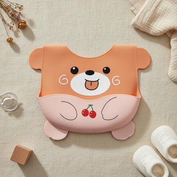 Orange bear cherry silicone baby bib flat lay showing full burnt orange bear shaped bib with round ears chubby arms bear feet expressive face tongue out and blush pink tummy pocket with red cherry illustration on linen background with wooden block white pacifier and white baby socks