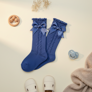 Steel blue satin bow knee-high baby girls socks – premium flat lay on cream background with wooden rattle, white baby shoes and pacifier showing cable knit texture and satin bow detail