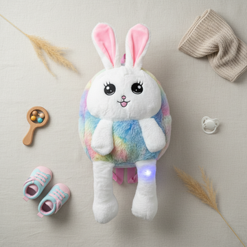 Rainbow bunny LED light-up plush backpack – lifestyle flat lay with baby shoes, wooden rattle and pacifier showing full rainbow body