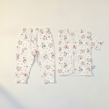 Happy Cosy Cartoon Monster Animal Print Baby Pyjama Set - premium studio lifestyle flat lay on beige linen showing long sleeve top and trousers with wooden elephant teether white baby shoes and dried lavender