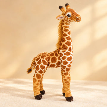Realistic standing giraffe plush toy studio scene on warm cream sand background with dappled natural sunlight showing three-quarter side view of life-like standing giraffe plush with deeply textured rust-brown and cream polygon patch coat golden mane along long neck dark ossicones large glassy eyes soft pink ears fluffy tufted tail and dark chocolate hooves in natural standing pose