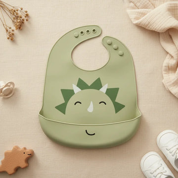Sage green dinosaur silicone baby bib flat lay showing all-sage-green bib with smiling triceratops face dark green spikes white horns closed happy eyes and matching sage green food catch pocket with embossed smile on linen background with wooden dinosaur teether white pacifier and white lace-up sneakers